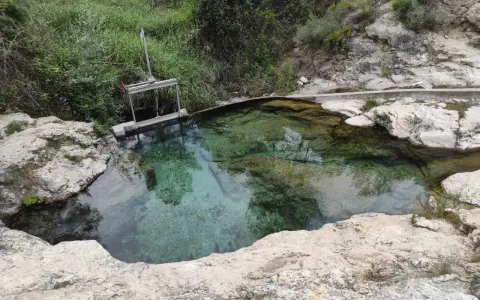 Aguas cristalinas en los baños de Somogil, rodeadas de rocas y vegetación. Una compuerta metálica controla el flujo del agua.
