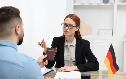 Kobieta w okularach przekazuje paszport mężczyźnie, omawiając zarobki w Niemczech. Flaga Niemiec na biurku.