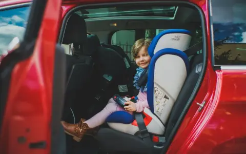Uśmiechnięta dziewczynka w foteliku samochodowym Britax Römer w czerwonym aucie.