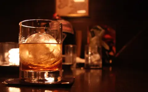 Ein Glas mit bernsteinfarbenem Whisky und einer großen Eiskugel steht auf einem dunklen Holztisch. Man trinkt Whisky mit Eis für den perfekten Genuss.