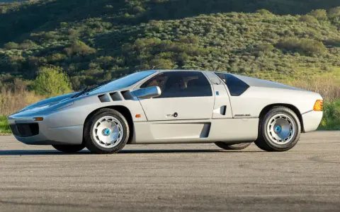 Silberner Isdera Imperator 108i mit markanten Lufteinlässen und futuristischem Design, geparkt vor einer grünen Hügellandschaft.