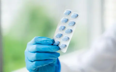 Ręka w niebieskiej rękawiczce trzyma blister z niebieskimi tabletami. To leki na obniżenie popędu seksualnego bez recepty, które mogą pomóc w rozwiązaniu problemów z erekcją.