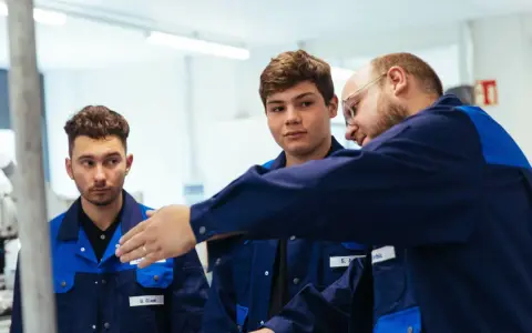 Drei junge Männer in blauen Arbeitskitteln lernen gemeinsam. Ein Ausbilder erklärt etwas, während die genz-Auszubildenden aufmerksam zuhören.