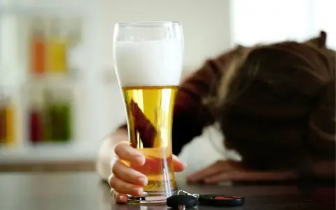 Ręka trzymająca piwo, obok kluczyki samochodowe. Osoba w tle pochylona.