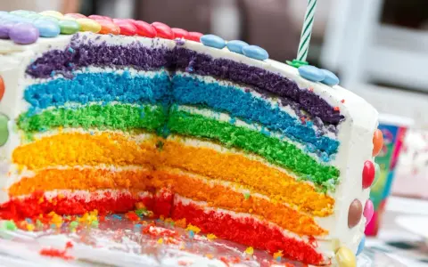 Kolorowy tort tęczowy z warstwami ciasta w kolorach tęczy, ozdobiony cukierkami i świeczką.