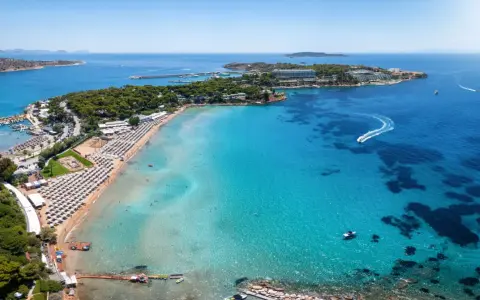 Traumhafter Blick auf das athen meer mit einer belebten Strandpromenade, Liegestühlen und dem glitzernden, türkisfarbenen Wasser, auf dem Boote fahren.