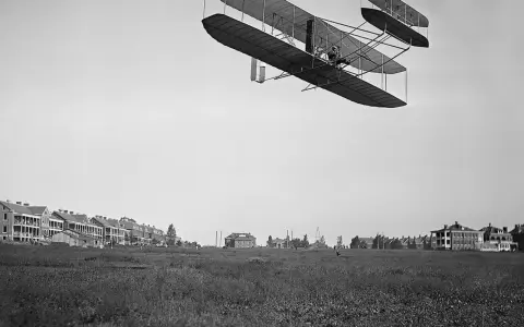 Das Flugzeug der Gebrüder Wright fliegt über ein Feld. Im Hintergrund sind Gebäude zu sehen.