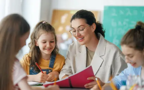 Lehrerin erklärt Kindern etwas aus einem Buch. Eine Tafel mit Rechenaufgaben im Hintergrund deutet auf die Bedeutung von Bildungspolitik für die Zukunft hin.