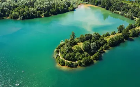 Türkisblaues Wasser umgibt eine grüne Insel mit einem Pfad am Niedernberger See. Ein Stand-Up-Paddler genießt die Ruhe.