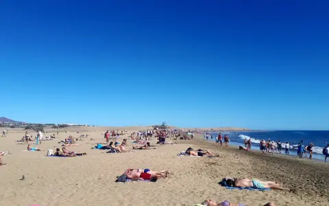 Tłum ludzi odpoczywa na słonecznej plaży. Woda jest spokojna, a niebo bezchmurne. Czy Gran Canaria jest bezpieczna? Tak, wygląda na to, że turyści czują się tu swobodnie.