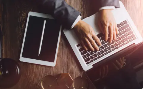 Dłonie na klawiaturze laptopa, obok tablet i młotek sędziowski. Profesjonalne narzędzia do pracy, być może związane z **taxat windykacja**.