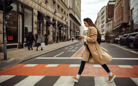 Kobieta w beżowym płaszczu i żółtych sneakersach przechodzi przez pasy. Idealne buty do trencza na miejski spacer.