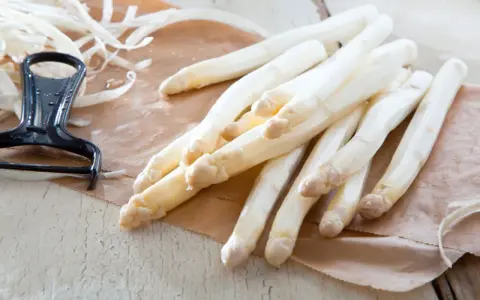 Frischer weißer Spargel liegt auf braunem Papier, daneben ein Sparschäler. Die Frage, warum riecht Urin nach Spargel, beschäftigt viele.