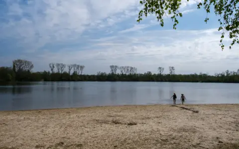 Zwei Kinder planschen im Wasser am Sandstrand des Dieksees in Lingen. Bäume säumen das Ufer unter einem bewölkten Himmel.