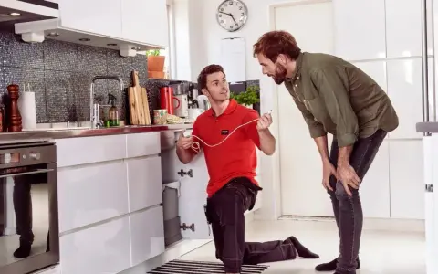 Tekniker installerar skalskydd larm under diskbänken i ett modernt kök. En man tittar på.