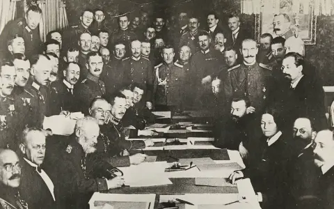 Delegierte unterzeichnen den Frieden von Brest-Litowsk. Männer in Uniformen und Anzügen sitzen an einem Tisch, umgeben von weiteren Personen.