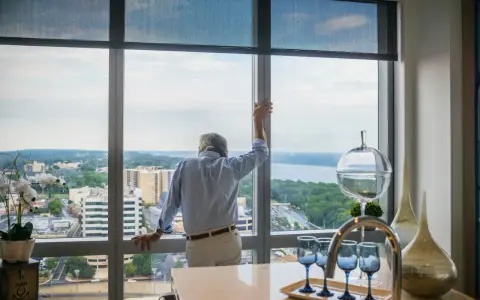 Hombre mira por ventana de aluminio, pensando cuánto pesa una ventana de aluminio. Vista urbana y río.
