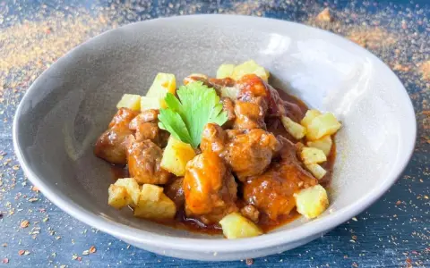 Delicioso cordero al chilindrón en un cuenco gris, acompañado de patatas fritas y una ramita de perejil.