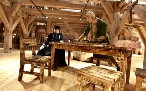 W muzeum Mazury dwie figury w strojach historycznych przy drewnianym stole. Jedna siedzi, druga stoi, pracując.