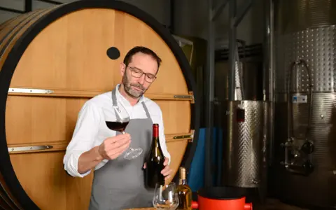 Un enologo esperto valuta un bicchiere di vino rosso, con una bottiglia in mano, davanti a una grande botte di legno e serbatoi d'acciaio.