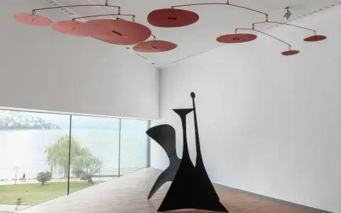 Un mobile Calder rouge flotte au-dessus d'une sculpture noire monumentale, dans une salle d'exposition lumineuse avec vue sur un lac.