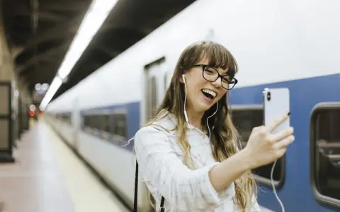 Junge Frau mit Kopfhörern macht ein Selfie am Bahnhof. Sie freut sich, ihr nächstes Abenteuer mit dem **discover eu pass** zu beginnen.