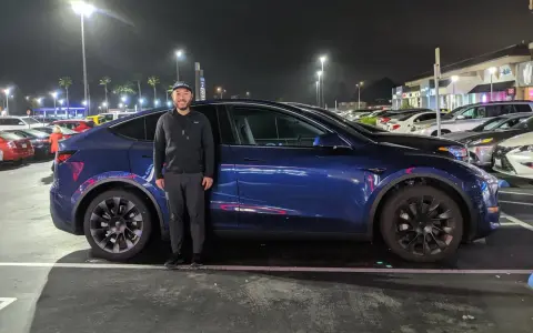 Mężczyzna stoi obok ciemnoniebieskiego Tesli Model Y na nocnym parkingu.