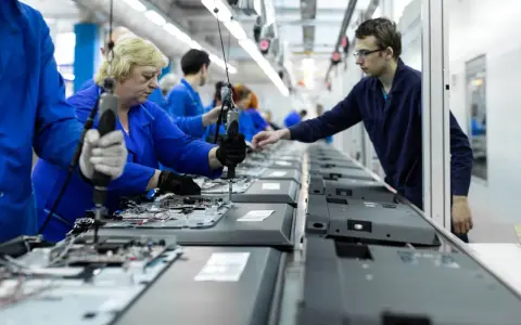 Pracownicy w niebieskich kombinezonach montują telewizory na linii produkcyjnej. To praca w ruchu ciągłym, gdzie każdy element jest kluczowy.