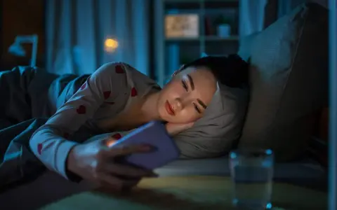 Une jeune femme dans son lit, le visage éclairé par son téléphone. Trop d'écrans, conséquences sur le sommeil.