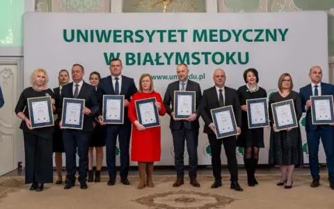 Grupa osób w garniturach i sukienkach pozuje do zdjęcia z dyplomami przed banerem Uniwersytetu Medycznego w Białymstoku.