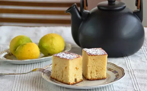Dos porciones de bizcocho de limón espolvoreadas con azúcar glas, junto a limones frescos y una tetera negra.