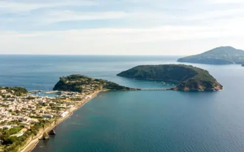 Panoramica di un'isoletta tra Napoli e Ischia, con un borgo costiero, un porto turistico e un ponte che collega a un'altra isola verdeggiante.