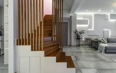 Nowoczesna, wysoka klatka schodowa aranżacje z drewnianymi stopniami i ażurową balustradą, wkomponowana w minimalistyczne wnętrze z szarą sofą i geometryczną ścianą.