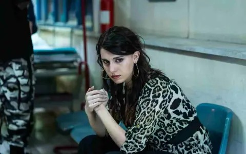 Carmela, con sguardo intenso, attende il suo destino fuori dal carcere. Indossa una camicia leopardata.