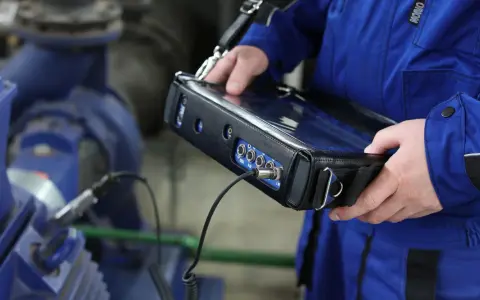 Technik w niebieskim kombinezonie wykonuje pomiary drgań maszyny przemysłowej za pomocą przenośnego urządzenia.