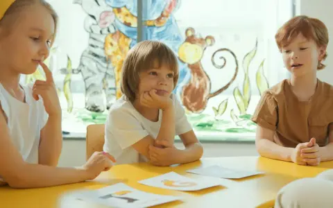Dzieci uczą się dodawania w zakresie 5, siedząc przy żółtym stole. Na kartkach widać obrazki zwierząt.