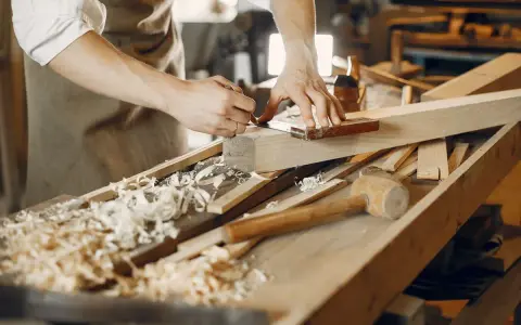Un artisan qui travaille le bois, concentré sur la précision de ses gestes. Des copeaux de bois jonchent l'établi.