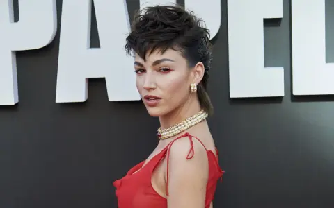 Mujer con un llamativo mullet largo, maquillaje rojo y vestido escarlata, posando ante un fondo con letras blancas.