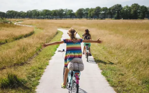 Zwei Mädchen genießen ein sonniges Fahrrad-Wochenende auf einem Feldweg, die Arme weit ausgebreitet.