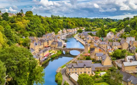 A picturesque view of Dinan, France, with its charming stone buildings lining a winding river, a stone bridge, and lush green hills under a cloudy sky.