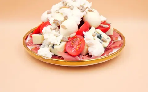 Wykwintna sałatka z szynką parmeńską, serami pleśniowymi, mozzarellą, pomidorkami i słonecznikiem na złotym talerzu.