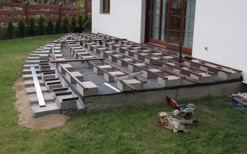 Rozpoczyna się montaż desek tarasowych na legarach. Podstawa z bloczków betonowych i podsypki piaskowej czeka na wykończenie.