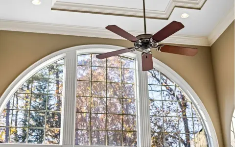 Ventilador de techo de madera oscura sobre una ventana arqueada con vista a árboles otoñales.