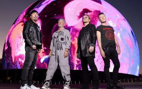 U2 w nowej płycie. Czterech członków zespołu stoi przed gigantycznym, kolorowym ekranem.