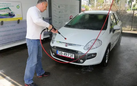 Mężczyzna myje białe auto na myjni. Na tablicy informacyjnej widnieje napis "Możliwość otrzymania faktury", sugerujący, że na myjni można płacić kartą.