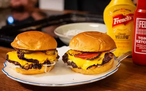 Två saftiga hamburgare med smält ost, pickles och lök på en tallrik. Bredvid står flaskor med senap och ketchup.