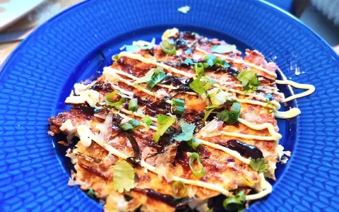 En läcker okonomiyaki på en blå tallrik, toppad med majonnäs, salladslök och torkad sjögräs. Ett perfekt okonomiyaki recept!