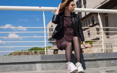 Dziewczyna w eleganckiej sukience i sportowych butach siedzi na deskorolce na schodach.