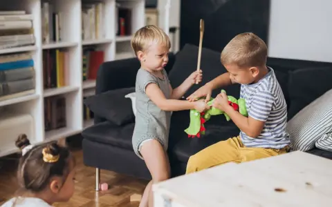 Zwei kleine Jungen streiten sich um ein grünes Stofftier. Einer hält einen Kochlöffel, der andere zieht am Spielzeug. Ein kleines Kind schaut zu.