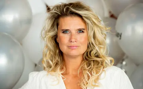 Blond kvinna med blå ögon och lockigt hår, omgiven av silverfärgade ballonger. Hon ser ut att vara redo för festliga udda veckor.